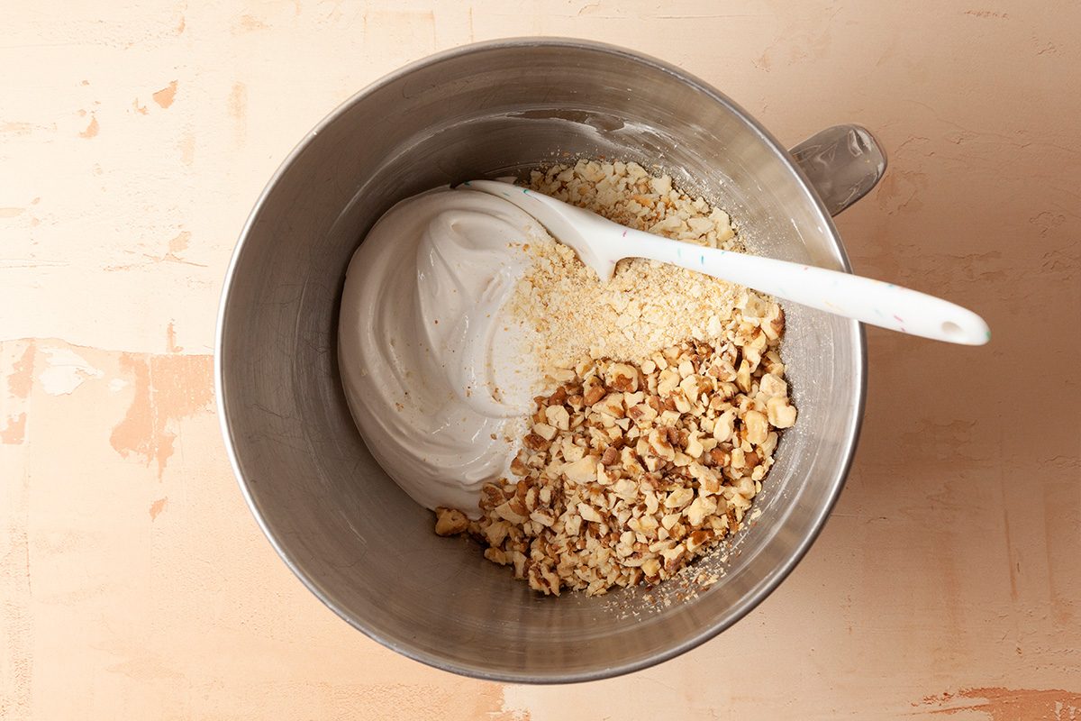 A metal mixing bowl containing whipped egg whites, chopped walnuts, and almond flour with a white spatula resting inside.