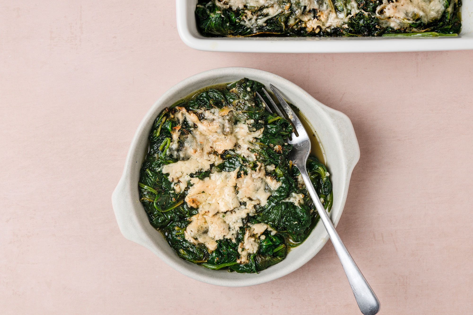 A top shot of a bowl of creamy baked spinach topped with golden melted cheese sits beside a fork; part of a baking dish appears above on a smooth light beige surface; the setting is cozy and appetizing