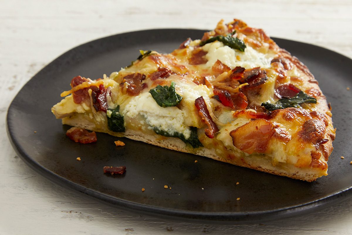 A slice of pizza on a dark plate, topped with melted cheese, spinach, ricotta, and pieces of crispy bacon.