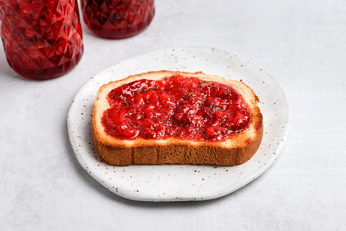 A slice of bread topped with strawberry jam sits on a white plate, with jars of jam visible in the background.