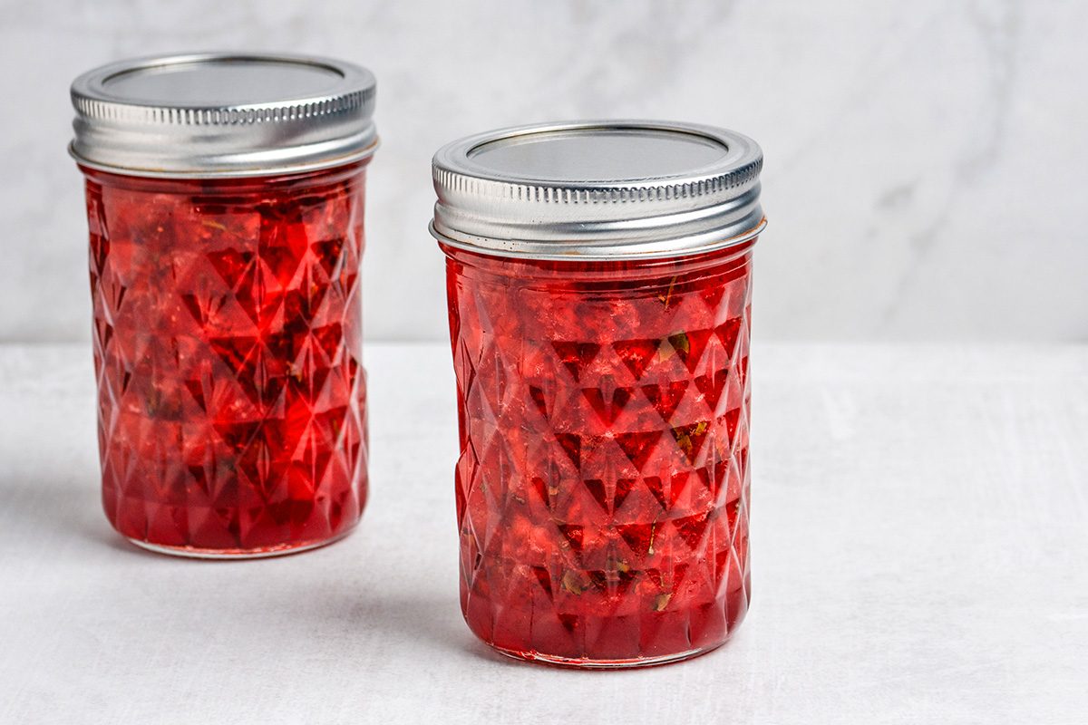 Two glass jars filled with bright red jam or jelly, sealed with silver metal lids, are placed on a white surface with a light gray background.