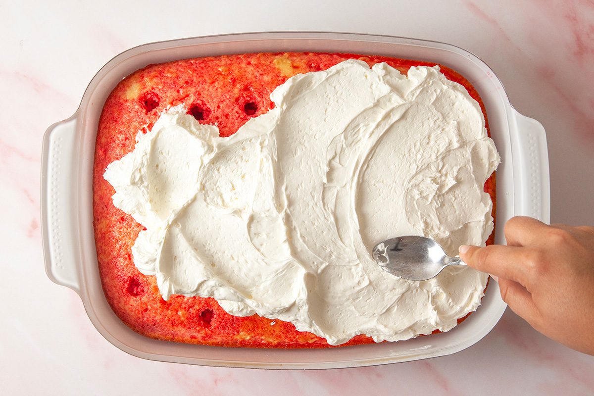 overhead shot of a hand spreads white whipped topping over a rectangular strawberry poke cake in a white baking dish, using a spoon; The cake has red spots where strawberry syrup was poured into hole