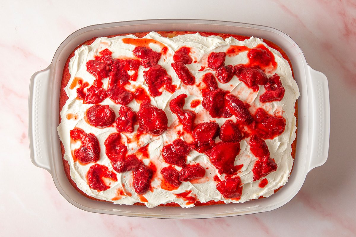 overhead shot of a rectangular baking dish filled with a dessert topped with whipped cream and drizzled with a chunky strawberry sauce, set on a light pink marble surface