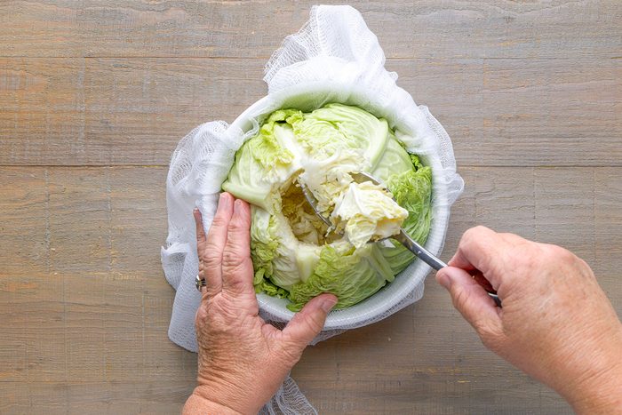 A person scooping out the center of a head of cabbage with a spoon, while holding it steady on a wooden surface covered with white cloth.