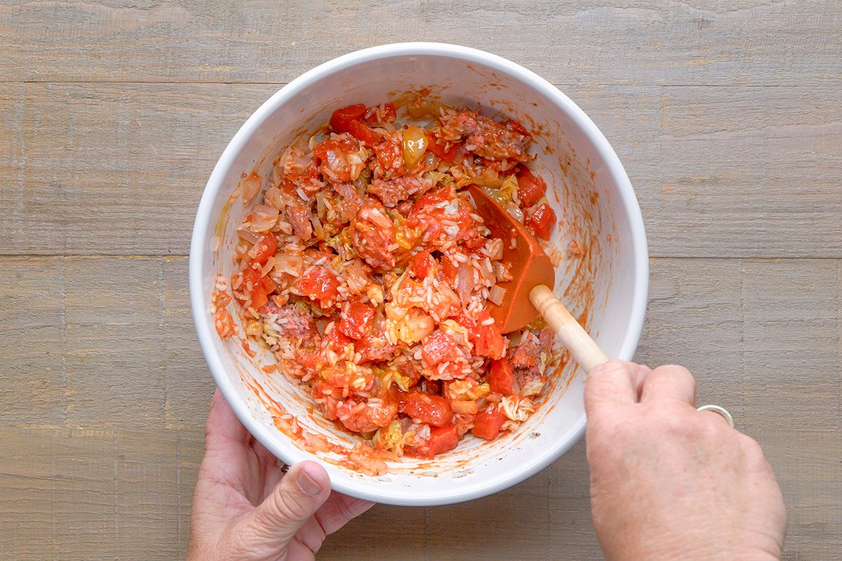 A person stirs a mixture of chopped tomatoes, rice, and seasonings with an orange spatula in a white bowl on a wooden surface.