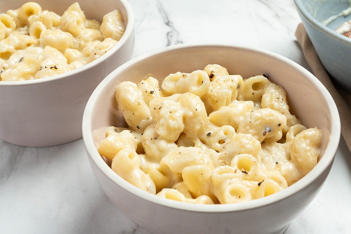 Two white bowls filled with creamy macaroni and cheese sit on a marble surface. The pasta is coated in a rich, cheesy sauce and topped with a sprinkle of black pepper.