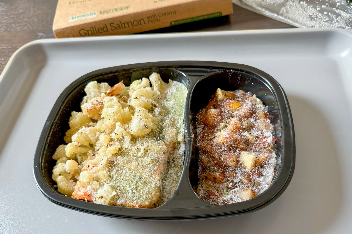 A frozen meal in a black plastic tray sits on a gray surface. The tray is divided into two sections: one with pasta and the other with a meat and vegetable mix, both covered in frost. A cardboard box is visible in the background.