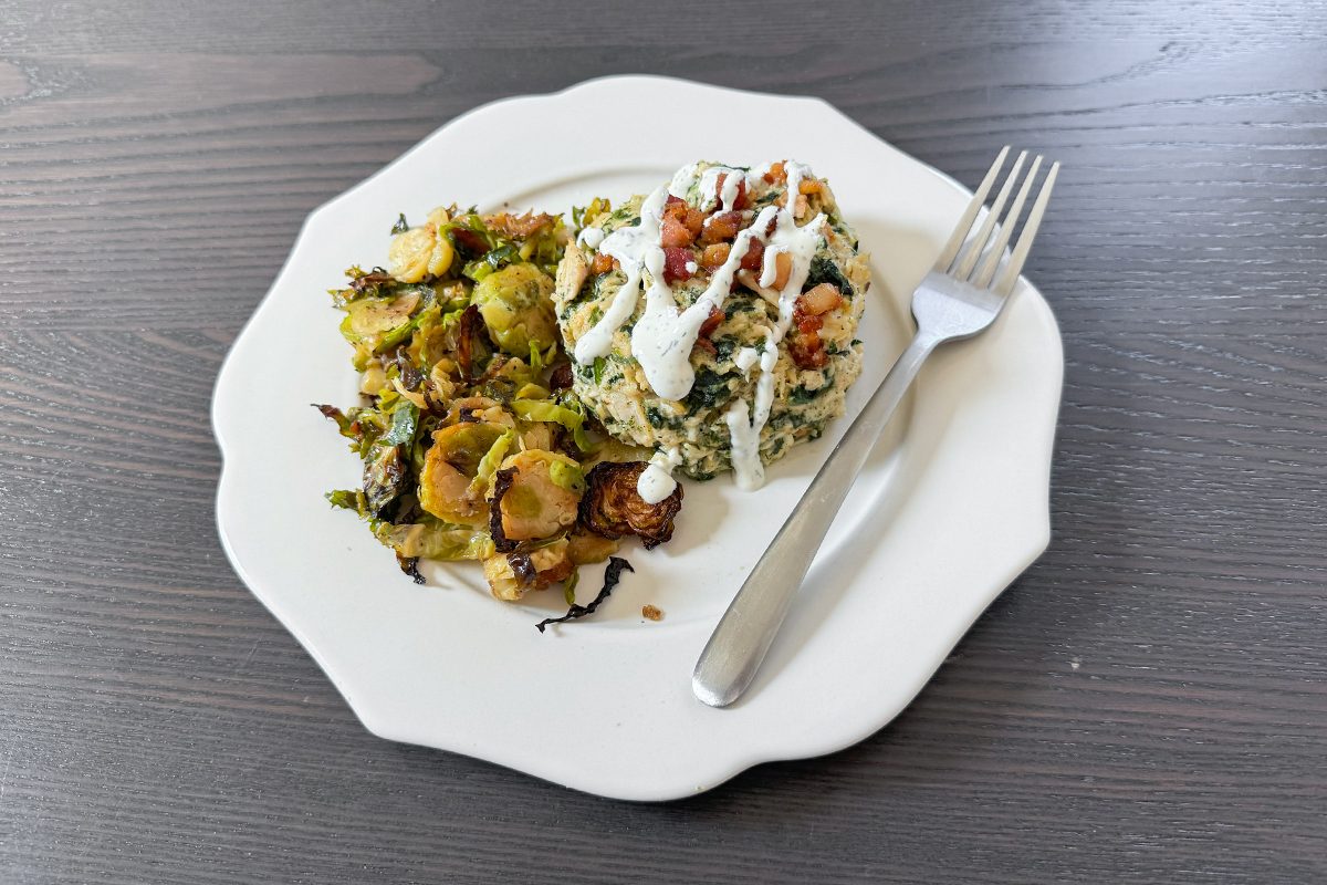A white plate with roasted Brussels sprouts and a serving of rice topped with sauce and nuts, next to a fork on a dark wooden table.