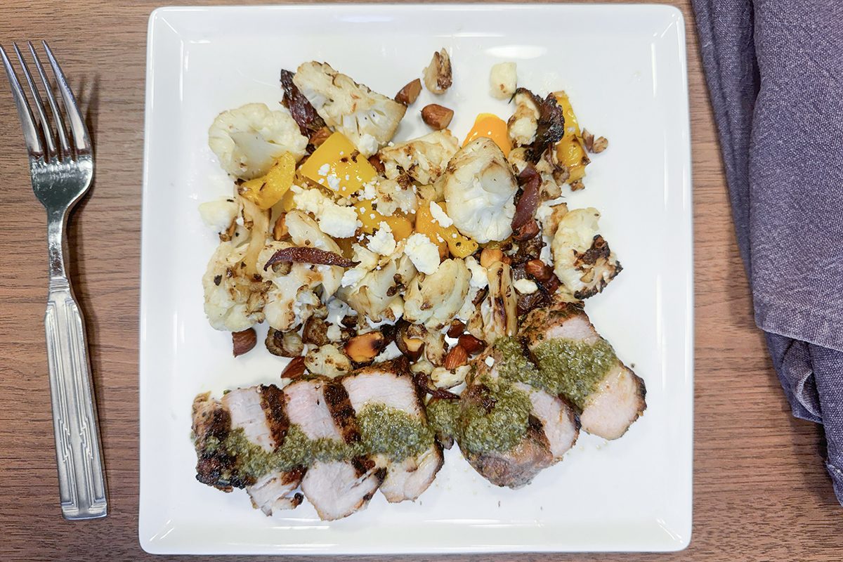 A square white plate with sliced pork topped with green pesto, served beside roasted cauliflower, yellow bell pepper, nuts, and cheese crumbles. A fork sits to the left and a folded purple napkin is on the right.