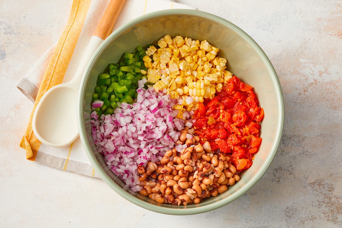 A bowl filled with chopped green bell pepper, corn, diced tomatoes, red onion, and black-eyed peas, arranged in sections. A white spoon and kitchen towel are beside the bowl on a light surface.