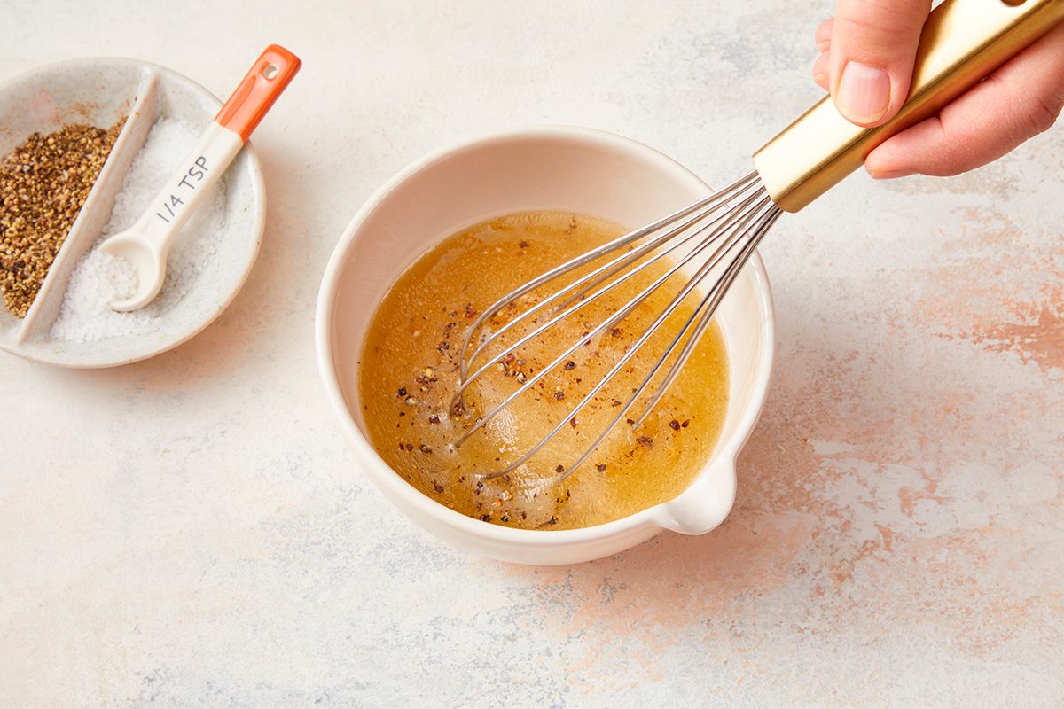 A hand whisks a yellow liquid with black pepper in a white bowl. Next to the bowl, there is a plate with dried herbs and a measuring spoon labeled “1/2 tsp.”.