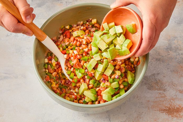 A person adds diced avocado from a small bowl into a larger bowl filled with a colorful bean and vegetable salad, mixing with a wooden spoon.