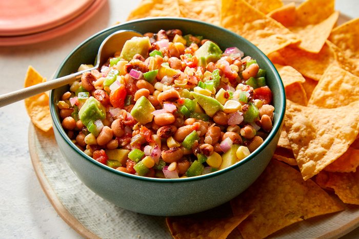 A bowl of colorful bean salad with chopped vegetables sits on a plate, surrounded by crispy tortilla chips. A spoon rests in the bowl, ready for serving.
