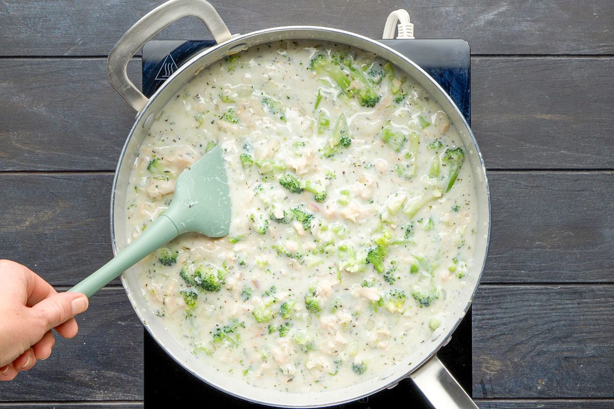 Top Shot of a hand stirs a thick; creamy broccoli and chicken mixture in a large skillet on a stovetop using a light green spatula; herbs and chopped broccoli pieces are visible in the sauce
