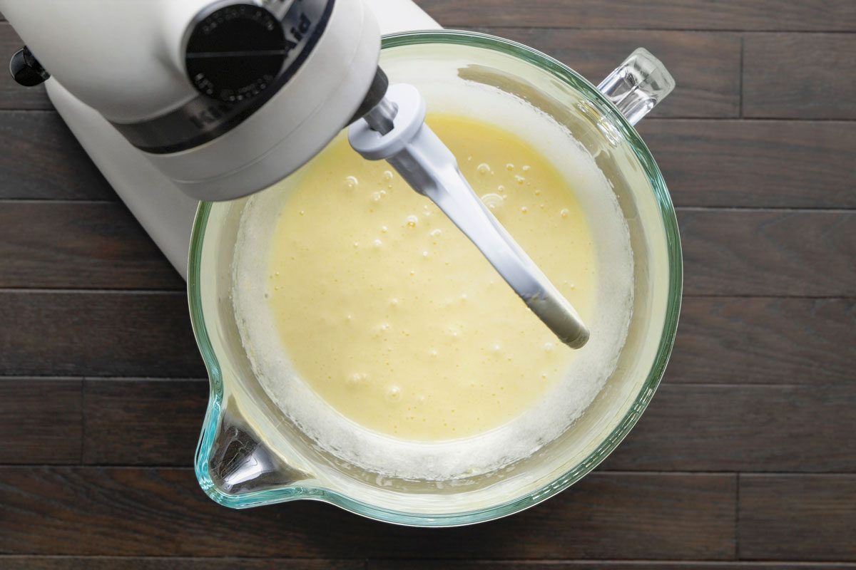 Overhead shot of a large bowl with eggs and sugar being beaten until frothy using a blender; all set on a dark brown wooden surface;