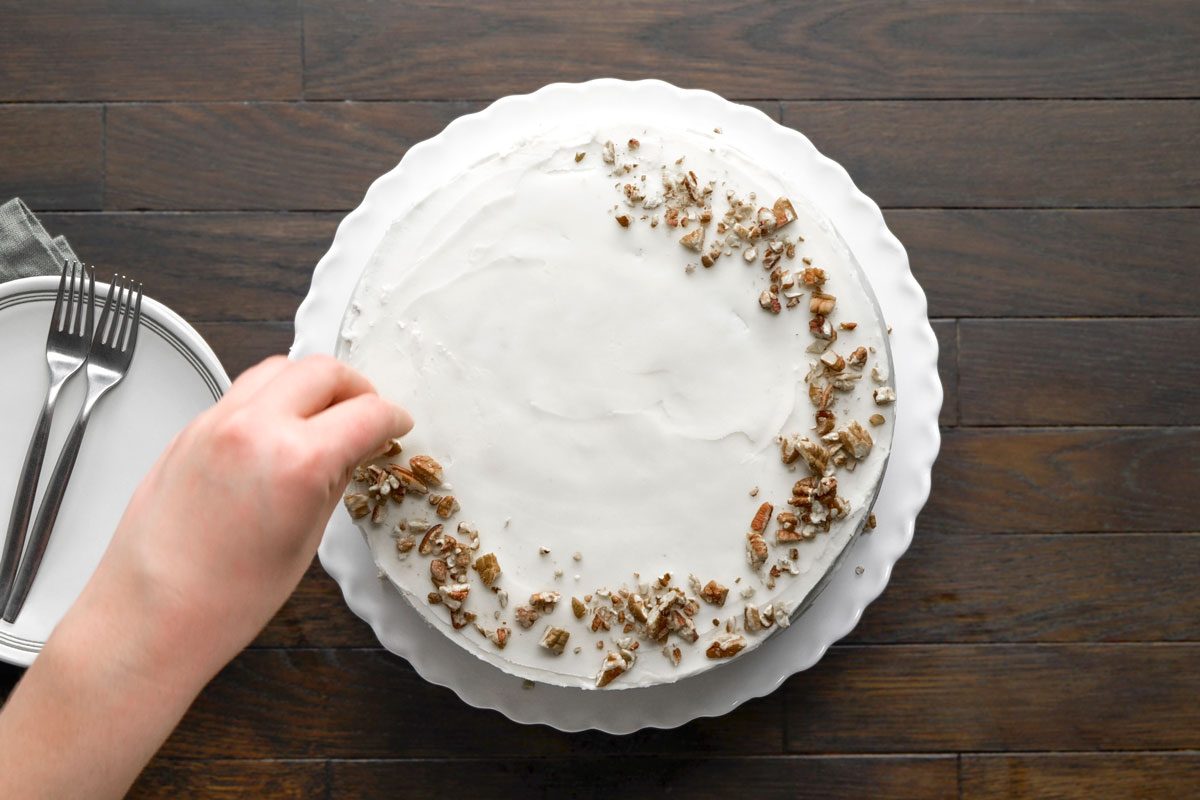 Overhead shot of Zucchini Carrot Cake; garnish with whole or chopped nuts; accompanied by two plates and two forks placed on a napkin; all arranged on a dark brown wooden surface;