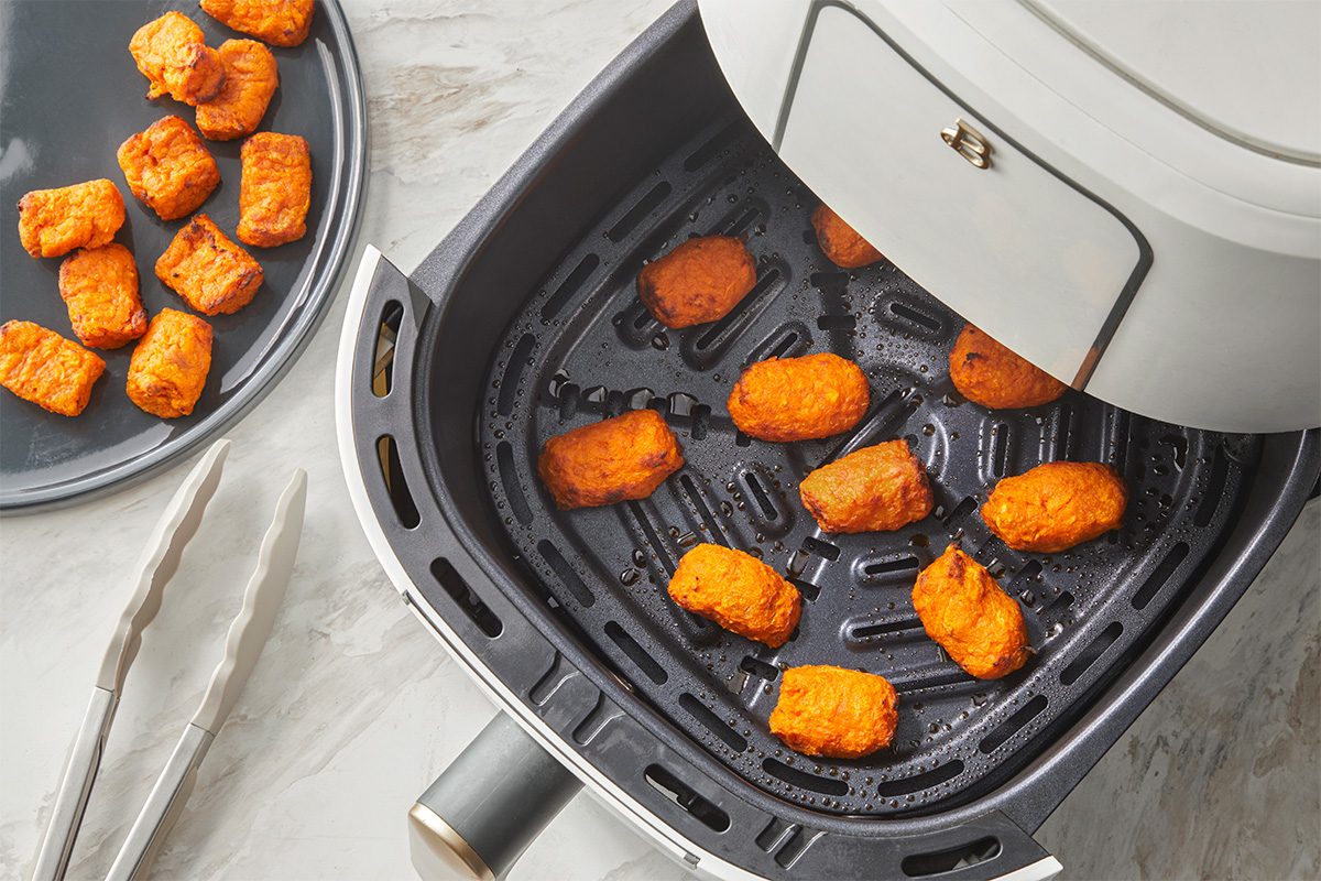 Golden brown nuggets are cooking inside an air fryer basket. A plate with a few finished nuggets and a pair of silver tongs are beside the air fryer on a light-colored countertop.