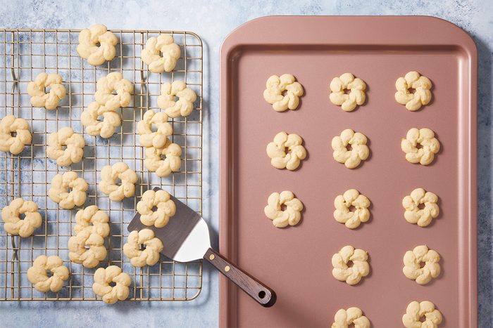 A pink baking sheet with unbaked flower-shaped cookies next to a cooling rack with baked cookies of the same shape. A spatula rests on the cooling rack. The background is a light blue surface.