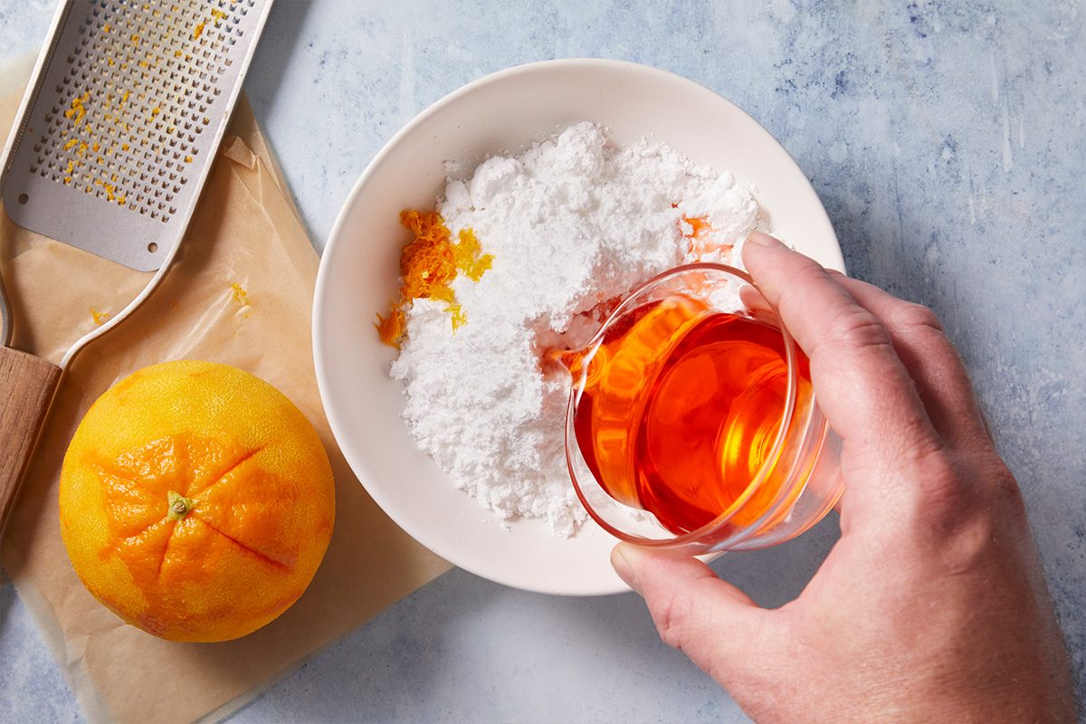 A hand holds a glass of orange liquid above a bowl containing powdered sugar and orange zest. A whole orange and a grater rest nearby on a light blue surface.