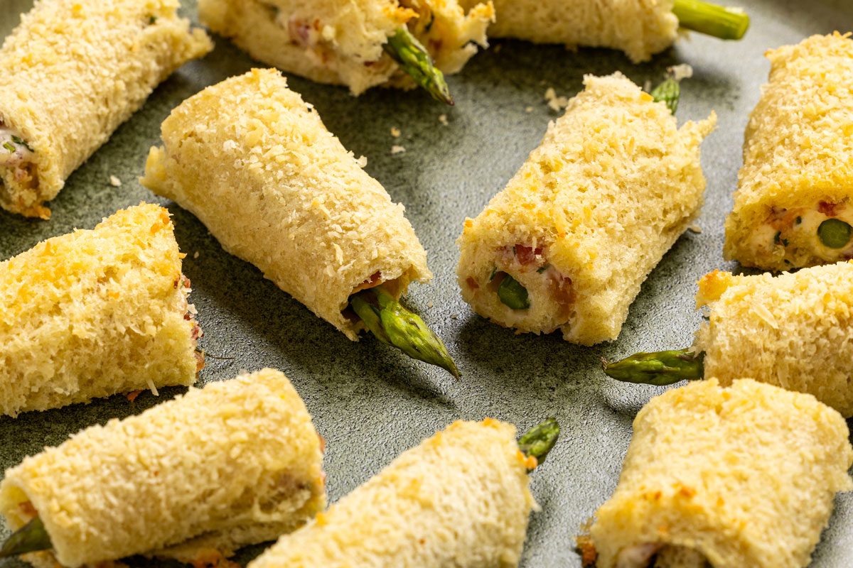 This is a top-down shot; multiple bread rolls stuffed with asparagus and creamy spread rest on a gray surface; bread appears toasted