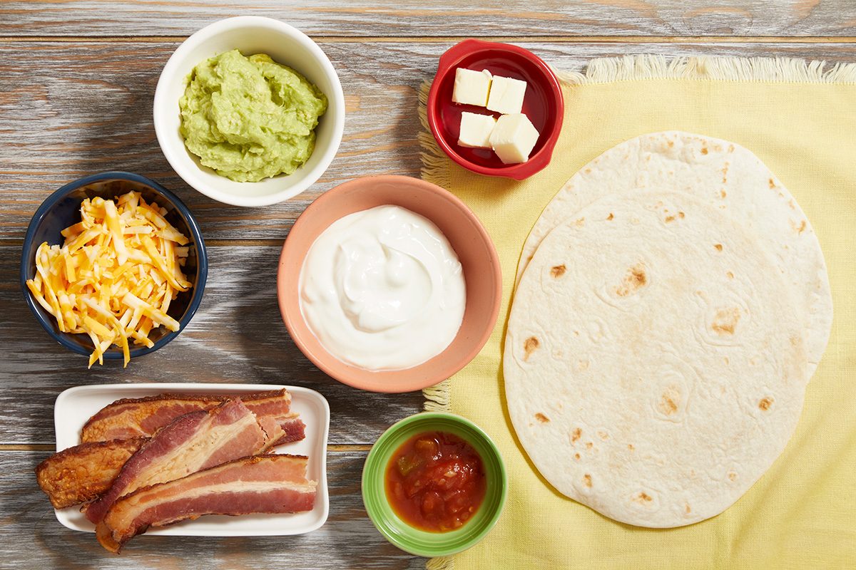 A top view of ingredients for making breakfast burritos, including flour tortillas, shredded cheese, guacamole, sour cream, salsa, diced cheese, and cooked bacon strips arranged on a wooden surface with a yellow cloth.