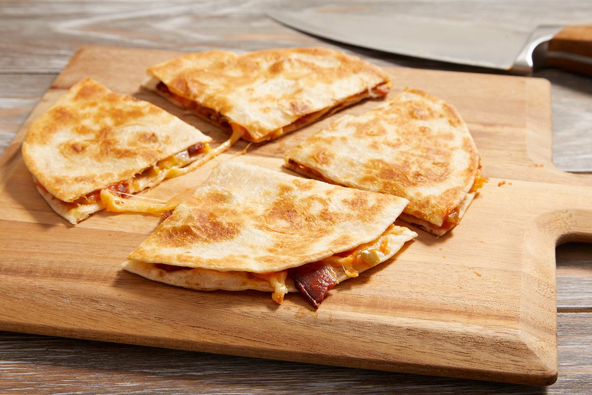 A quesadilla cut into four triangular slices sits on a wooden cutting board, with a knife nearby. The tortillas are golden brown and the filling appears to include melted cheese and meat.