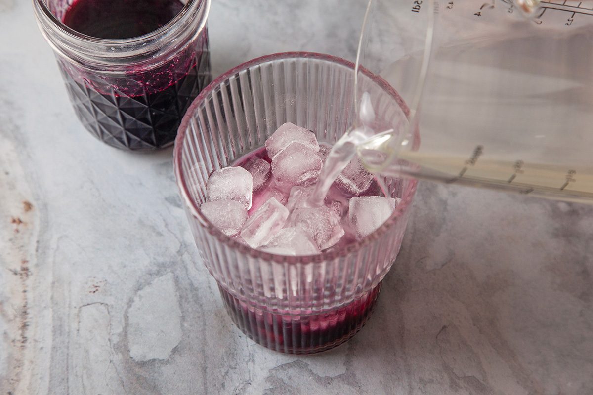 Overhead shot of a clear glass filled with ice cubes and purple liquid is being topped with a clear liquid; Nearby, there’s an open jar containing more purple liquid, all on a light-colored surface