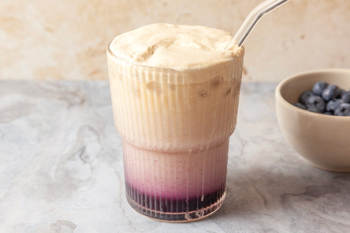 Close-up shot of a glass of Blueberry Cloud Coffee with a frothy top and a layer of purple syrup at the bottom; served with a glass straw; a bowl of blueberries is in the background