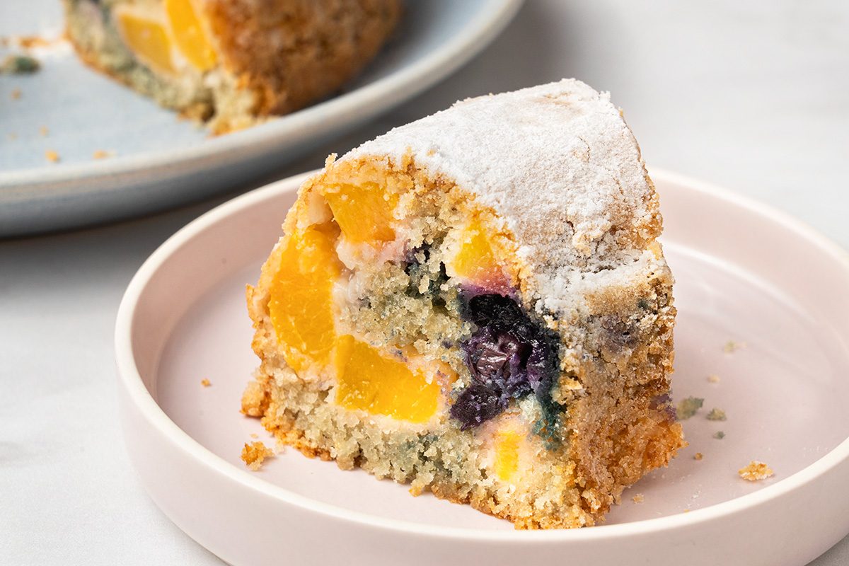 A slice of crumbly cake with visible pieces of yellow peach and dark blueberries, dusted with powdered sugar, served on a light pink plate.