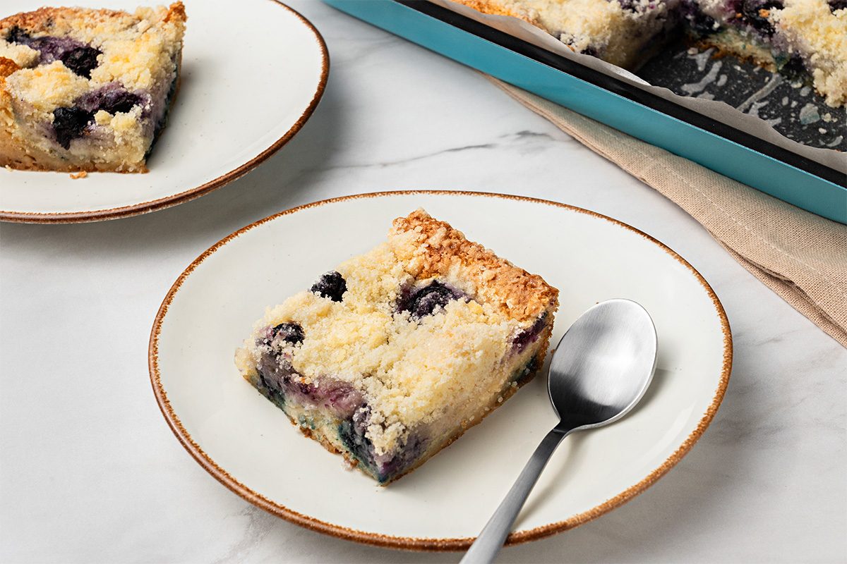 A square slice of blueberry crumb cake sits on a white plate with a spoon, with another plate and a baking dish of more cake in the background, all on a white marble surface.