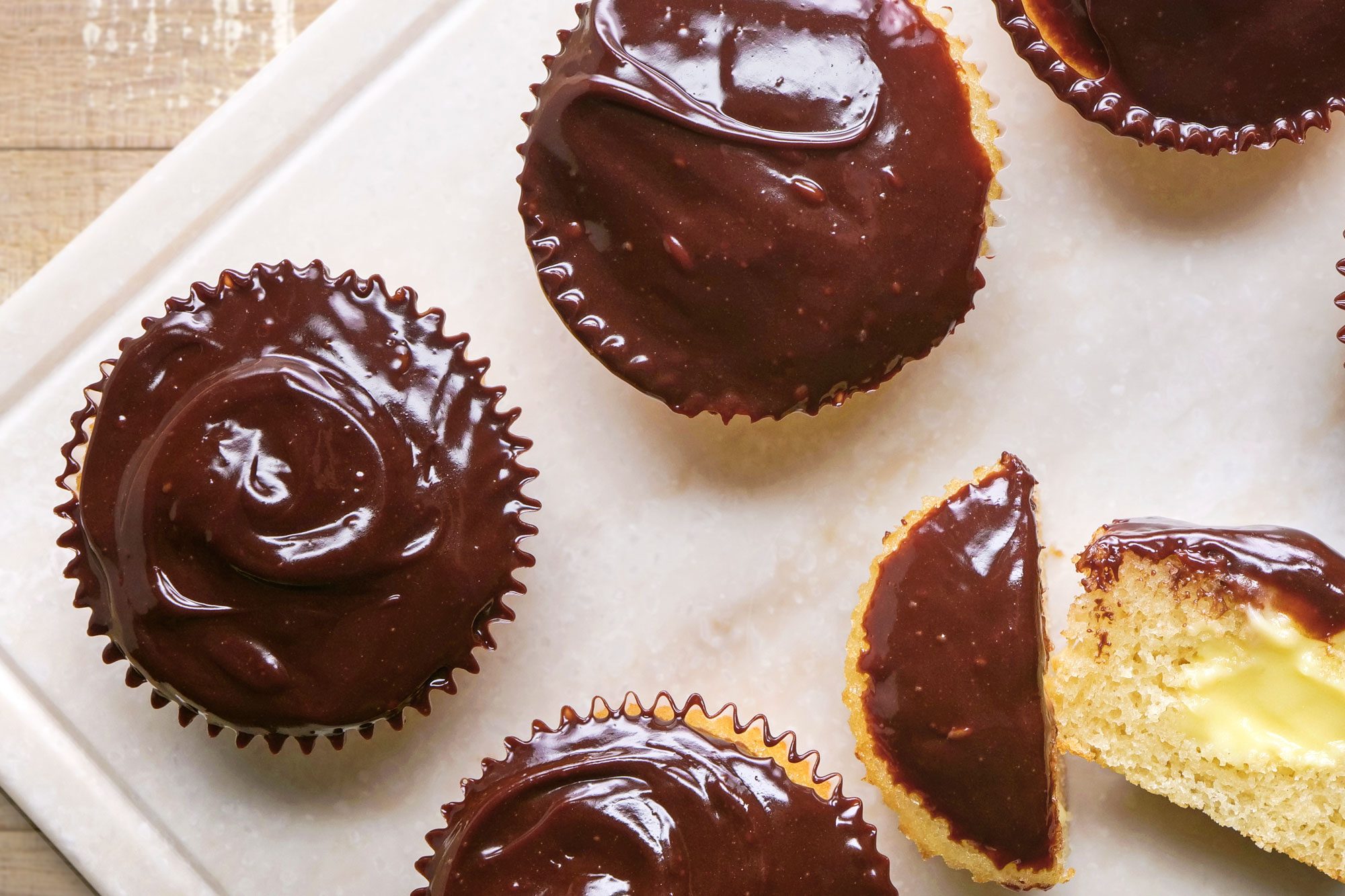 overhead shot of Six Boston Cream Cupcakes with glossy chocolate frosting are arranged on a white surface, with one cupcake cut in half to reveal a creamy yellow filling inside, The background is a light wooden surface