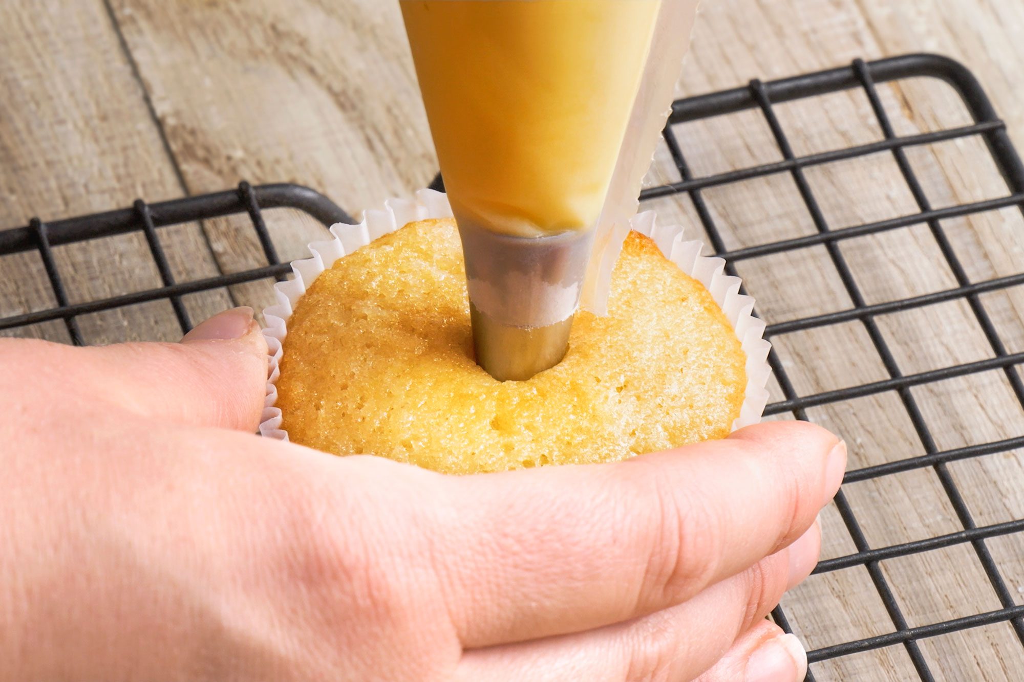 3/4th shot of hand holds a cupcake while a pastry bag filled with yellow cream is used to fill the center, with the cupcake resting on a black wire cooling rack on a wooden surface