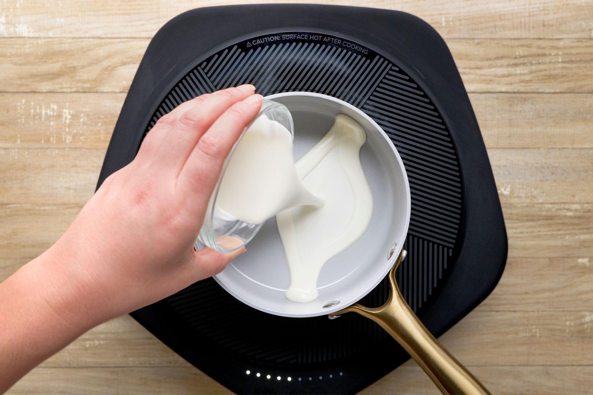 overhead shot of a hand pours milk from a small glass into a white saucepan with a gold handle, which is placed on a black stovetop over a light wood surface