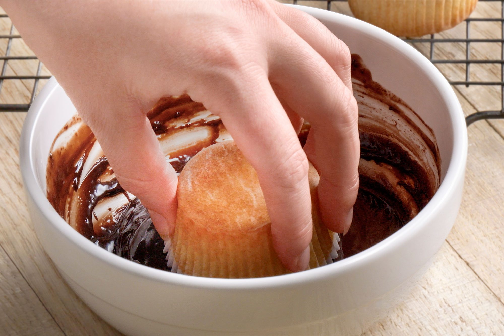 3/4th shot of a hand dips a plain cupcake into a bowl with chocolate frosting, preparing to coat the top, A cooling rack with another cupcake is visible in the background on a wooden surface