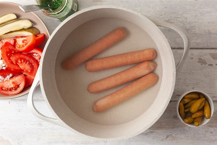 Four hot dogs boiling in a white pot of water on a light surface, with a plate of sliced tomatoes and pickles, a small bowl of pickles, and a drink visible nearby.