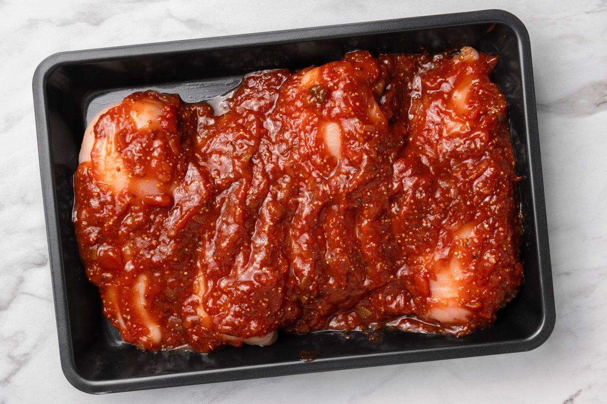Overhead shot of marinated chicken breasts in a baking sheet. Bake uncovered at 400 degree for 30–35 minutes; or until a thermometer reads 170 degree; all set on a marble surface;