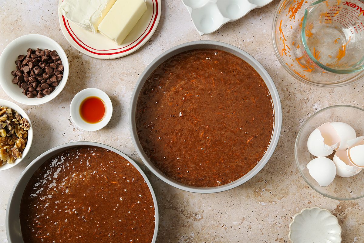 Two round cake pans filled with chocolate cake batter sit on a countertop surrounded by baking ingredients, including butter, cream cheese, eggs, chocolate chips, walnuts, vanilla extract, and a glass measuring cup.