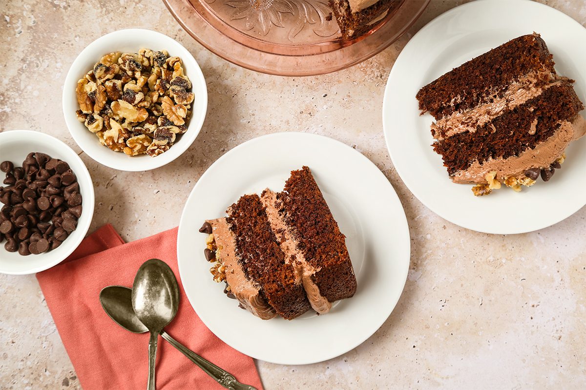 Two plates of chocolate layer cake with frosting and nuts, surrounded by bowls of chocolate chips and walnuts, a pink napkin, and two spoons on a light marble surface.