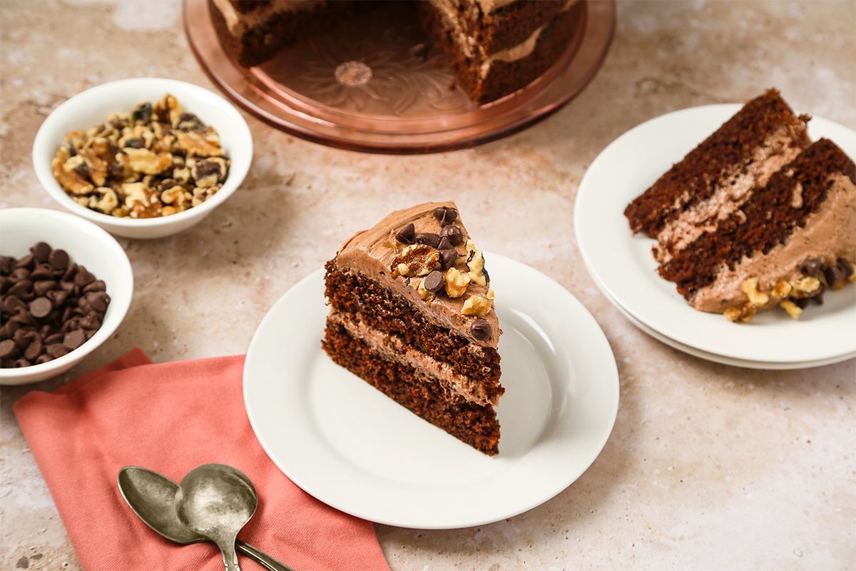 A slice of chocolate layer cake with chocolate frosting and walnuts sits on a white plate, next to a bowl of walnuts, a bowl of chocolate chips, two spoons, and a pink napkin. More cake is visible in the background.