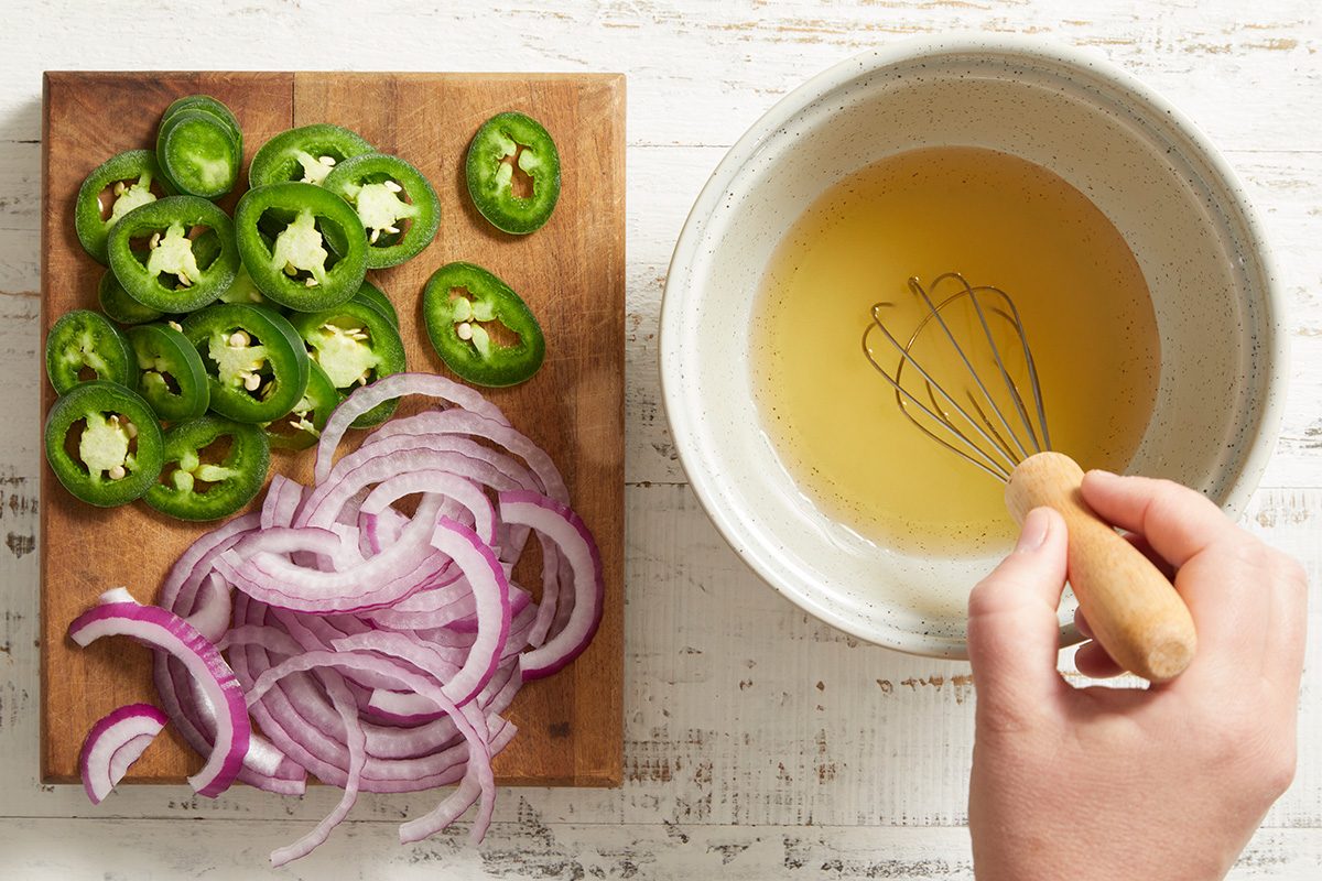 A hand whisks a yellow liquid in a white bowl next to a wooden board with sliced red onions and jalapeños on a white surface.