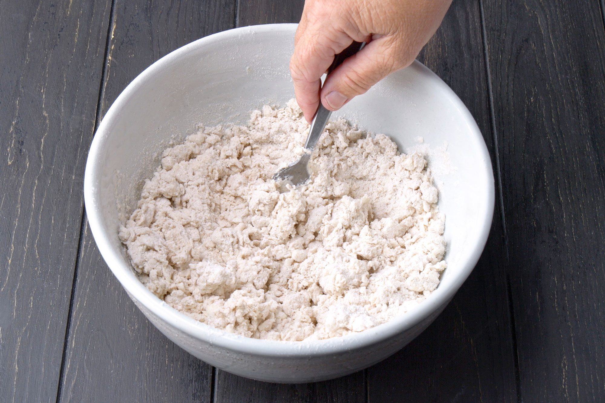 3/4th shot of a hand uses a fork to mix a crumbly dough mixture in a large white bowl on a dark wooden surface