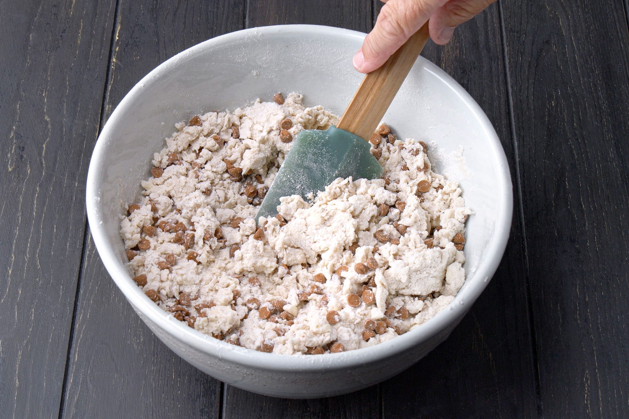 3/4th shot of A hand holds a spatula, mixing flour and cinnamon baking chips in a large gray bowl on a dark wooden surface