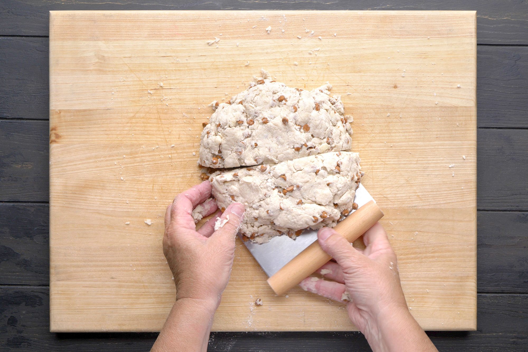 overhead shot of A person uses a bench scraper to cut a round ball of dough with nuts on a wooden cutting board; The board sits on a dark countertop