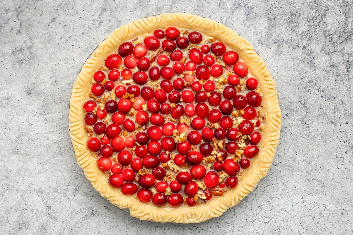 Unbaked pie with a crimped crust, filled with bright red cranberries and small pieces of pecans, on a gray textured surface.