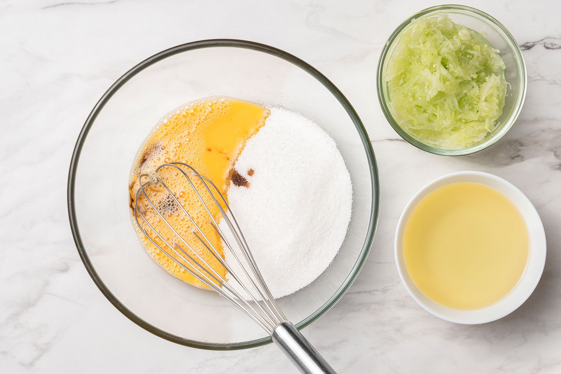 Sugar, oil, vanilla extract and grated, seeded and peeled cucumbers with eggs in bowls.