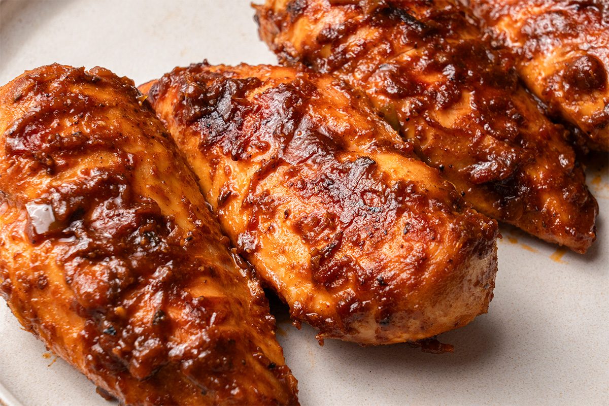 Three grilled chicken breasts coated in a rich, dark barbecue sauce are arranged on a light-colored plate. The chicken appears juicy with a slightly charred, caramelized exterior.