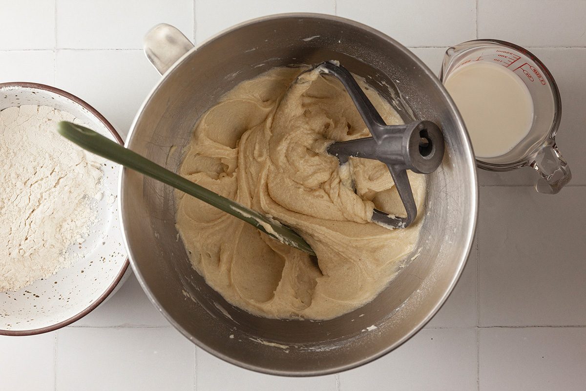 A mixing bowl with cookie dough, a green spatula, and a mixer attachment sits between a bowl of flour and a measuring cup of milk on a white surface.
