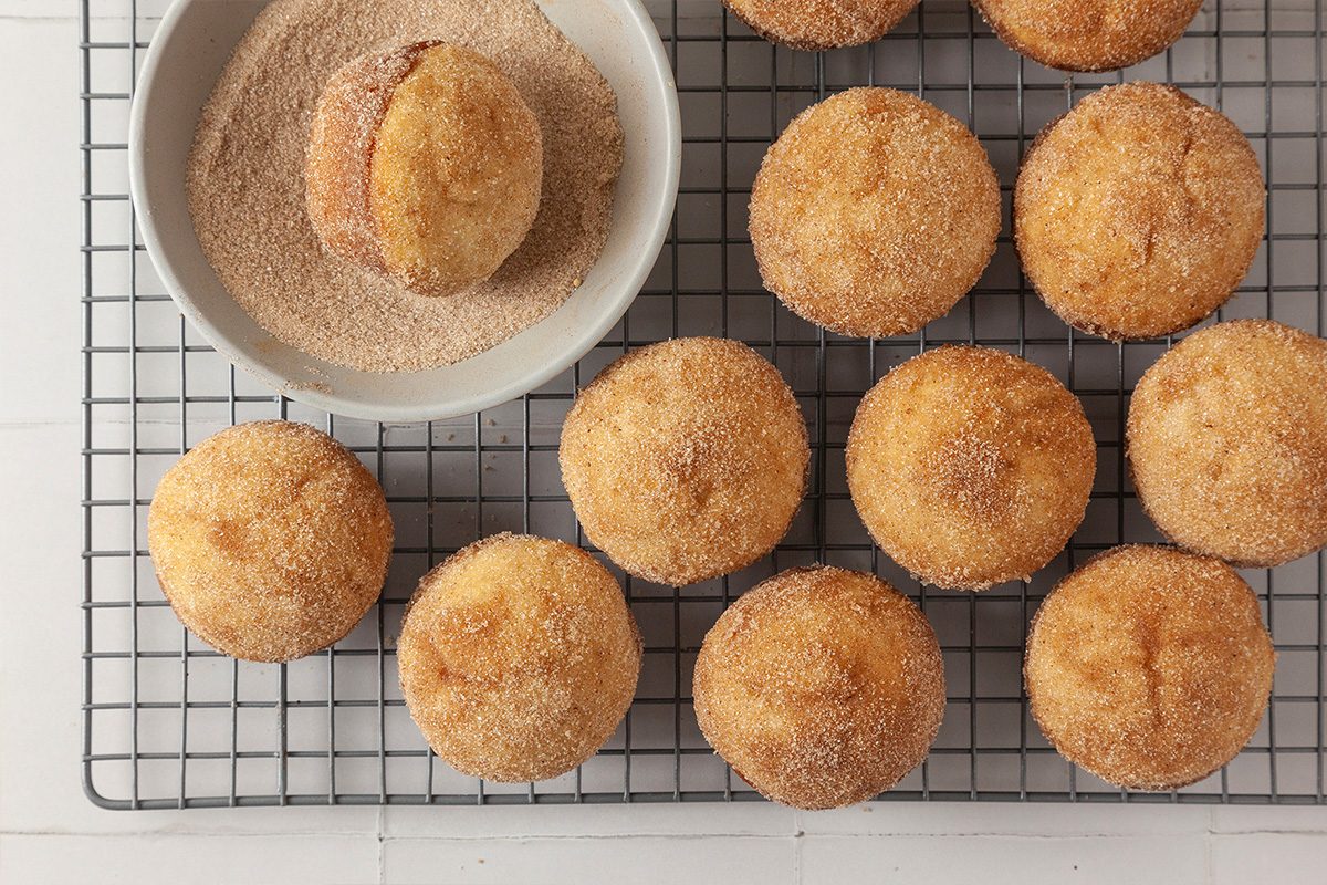 A wire cooling rack holds several round muffins coated with cinnamon sugar. One muffin is being rolled in a bowl of cinnamon sugar next to the rack. The background is a white tabletop.