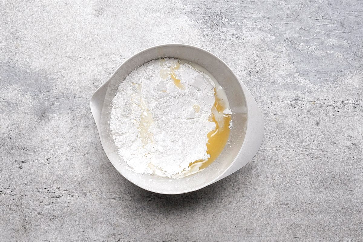 A white mixing bowl contains flour and a pool of yellow liquid, likely oil or melted butter, on a gray textured surface.