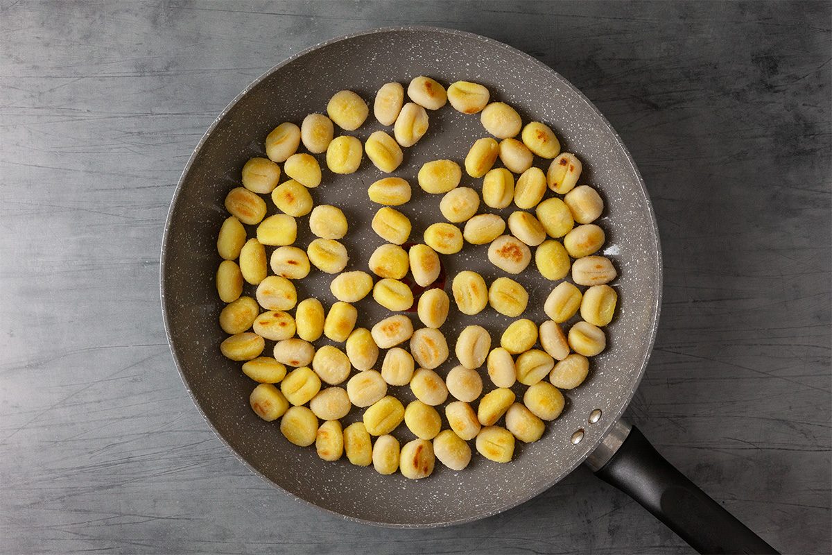 A nonstick frying pan filled with golden-brown gnocchi pieces sits on a gray surface. The gnocchi appear to be lightly sautéed or roasted, with some sides showing a toasted color.